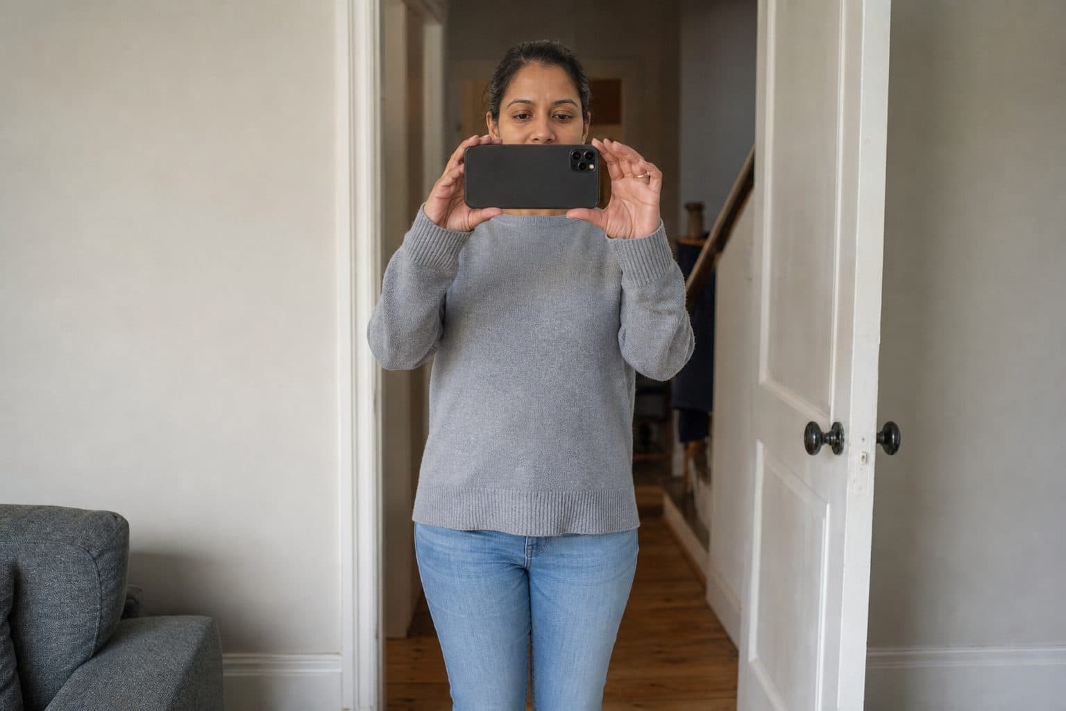 A woman standing in a doorway holds a phone sideways in both hands to photograph a living room, the back of the phone facing the camera